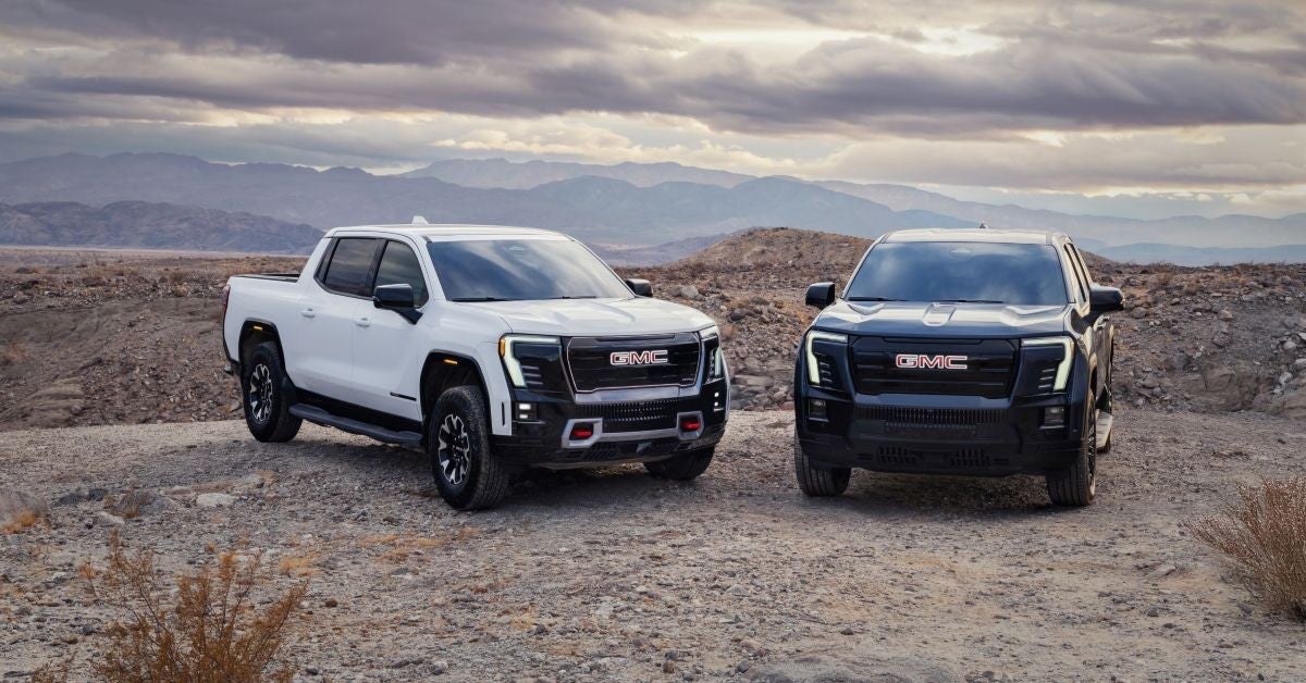 Two GMC pickup trucks, one white and one black, parked in a rocky desert with mountains in the background.