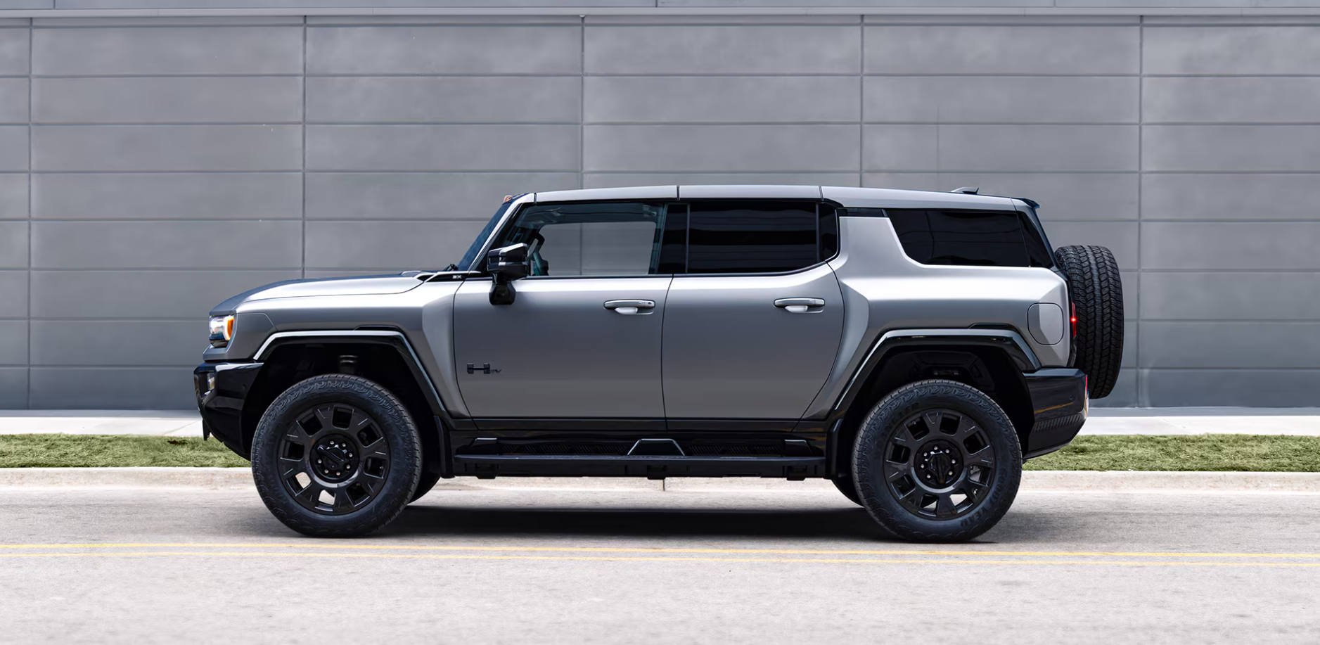 Side profile view of a silver 2026 GMC Hummer EV SUV parked on a city street in front of a modern gray building.